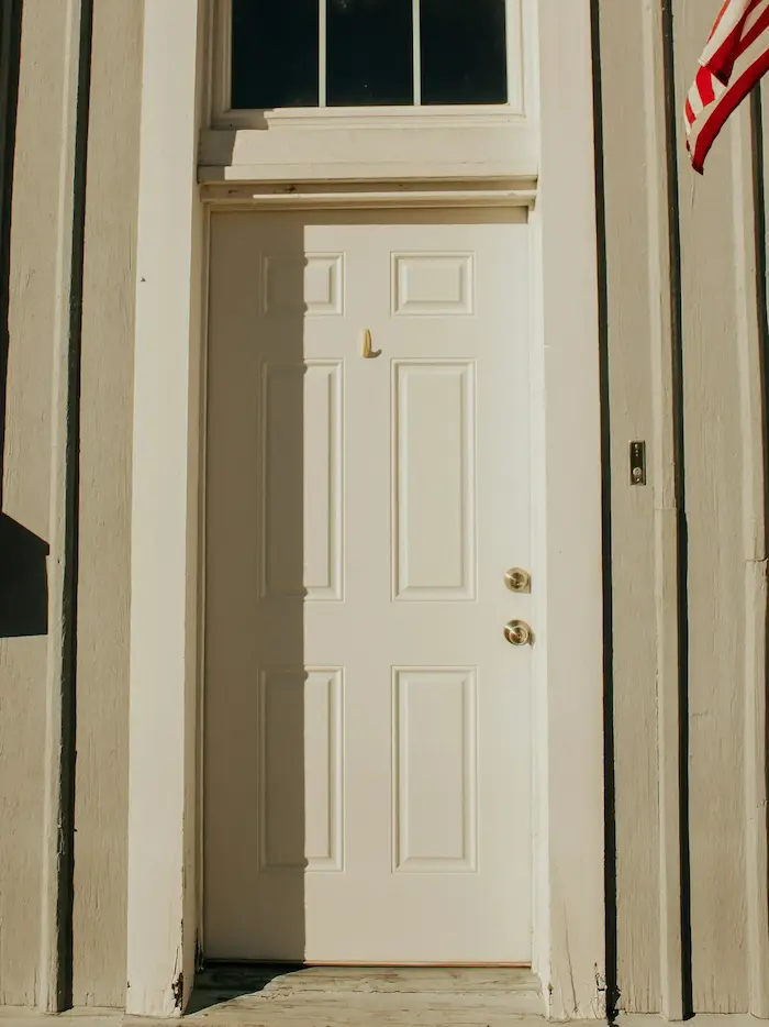 Une porte blanche avec une fenêtre en arc au-dessus, encadrée par des murs en bois peints en blanc et un drapeau américain visible sur le côté droit.