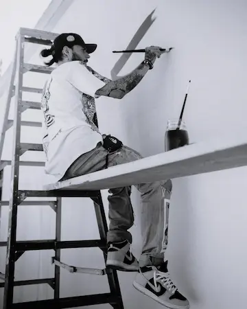 Un homme tatoué sur une échelle en train de peindre un mur blanc avec un pinceau, un pot de peinture posé à côté de lui.