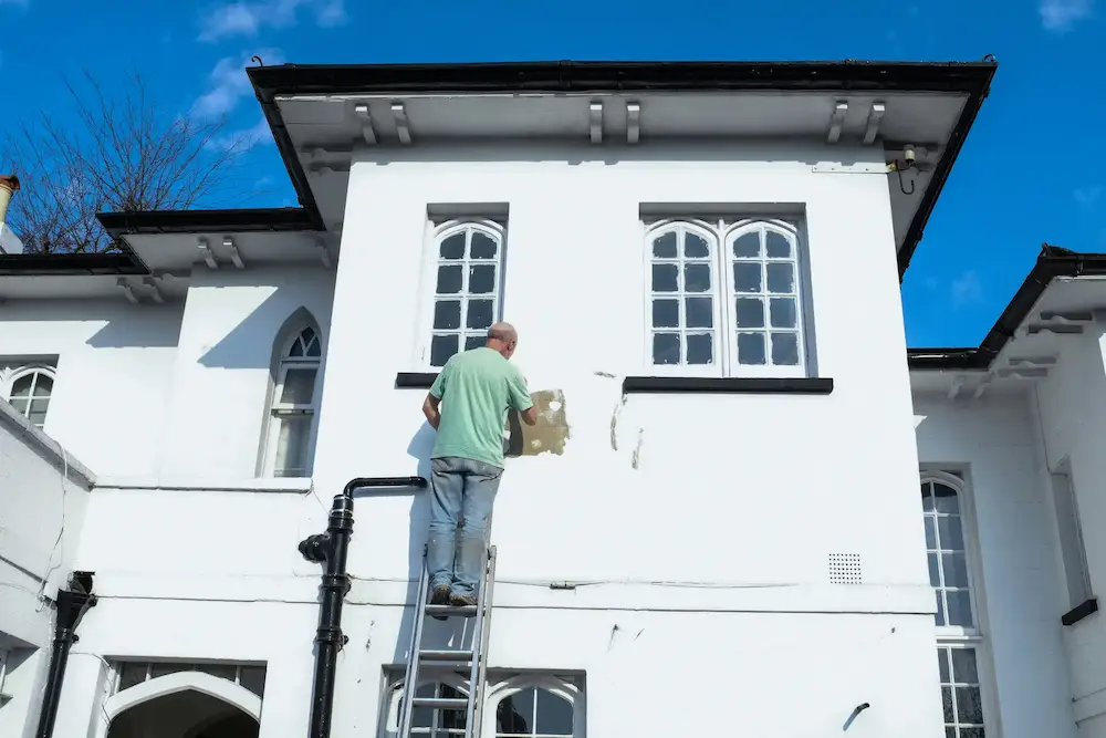 Un homme sur une échelle en train de peindre la façade blanche d'une maison avec des fenêtres à arcs, sous un ciel bleu clair.