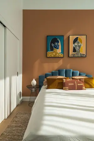 Une chambre à coucher avec un mur orange, un lit blanc avec des coussins colorés, deux tableaux abstraits au-dessus du lit, une table de chevet noire et un tapis en jute.