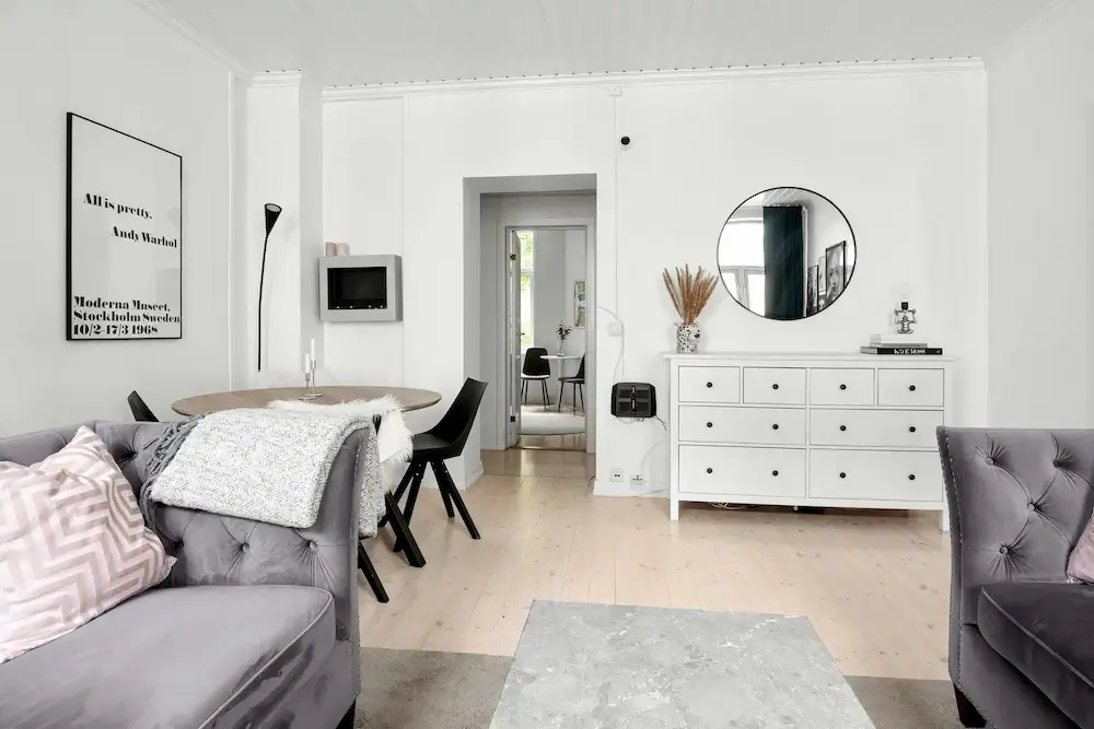 Un salon moderne avec un canapé gris, une table basse ronde, des chaises noires, un meuble blanc avec un miroir rond, et une affiche encadrée sur le mur.