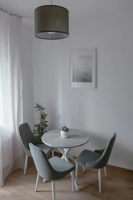 Un coin repas avec une table ronde blanche, trois chaises grises, une suspension noire au plafond, une plante verte sur la table, et un cadre accroché au mur.
