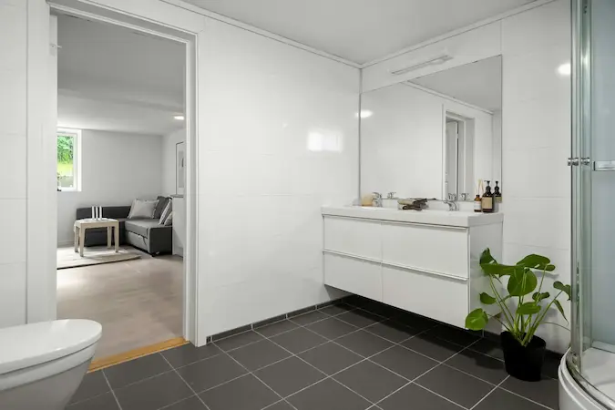 Une salle de bain moderne avec des murs carrelés en blanc, un sol en carrelage noir, un meuble de salle de bain blanc avec deux vasques, un grand miroir, et une plante verte dans un pot noir.