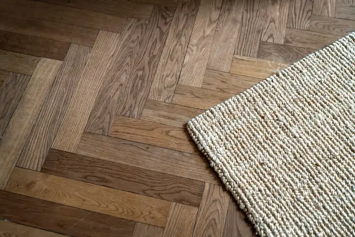 Un sol en parquet disposé en motif de chevrons, avec un tapis en laine tressée beige posé sur le côté droit.