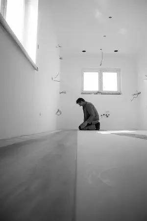 Un homme agenouillé dans une pièce en construction, avec des murs blancs, des fils électriques apparents, une fenêtre à deux battants, et un sol en béton brut.