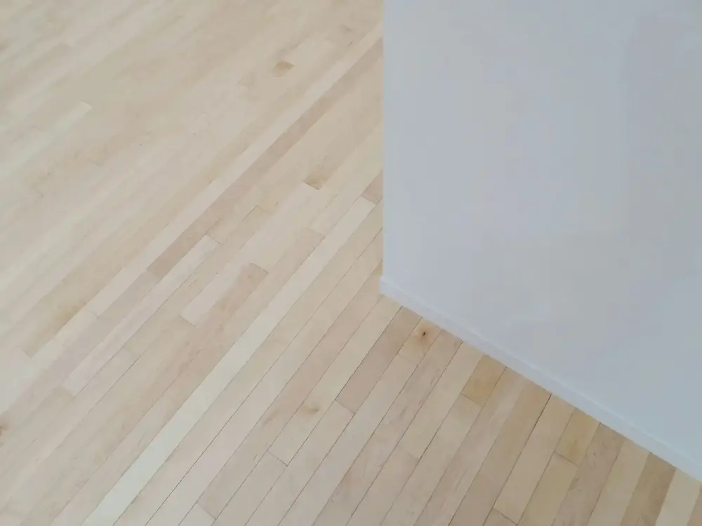 Un sol en bois clair avec des lattes disposées en diagonale, contrastant avec un mur blanc uni.