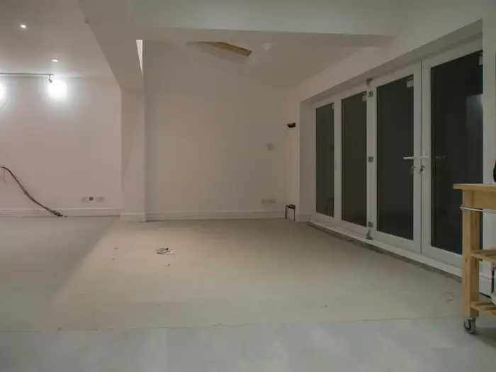 Une pièce vide en cours de rénovation avec des murs blancs, un sol en béton brut, une grande porte-fenêtre à trois battants sur le mur de droite, et des câbles électriques visibles sur le mur de gauche.